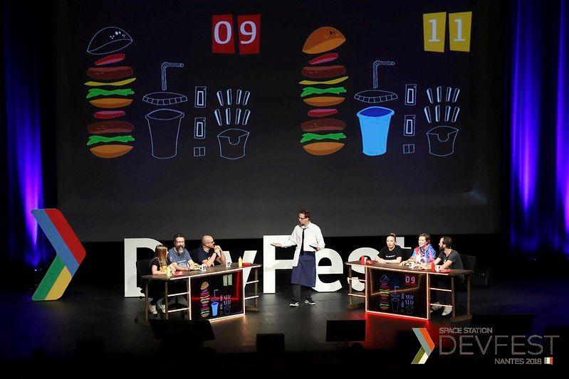 Image du burger quizz fait pendant le devfest nantes (écran avec les scores du burger quiz, 2 équipes de 3 par tables et un présentateur en tablier)