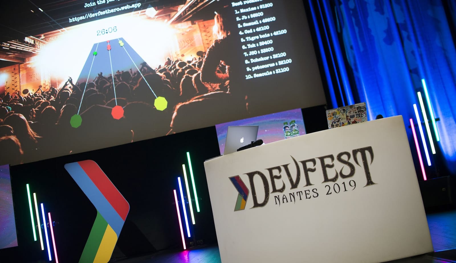 photo de la scène principale du Devfest Nantes 2019 (sur l'écran on voit un jeu type guitar hero et sur scène on voit un pupitre avec un ordi)